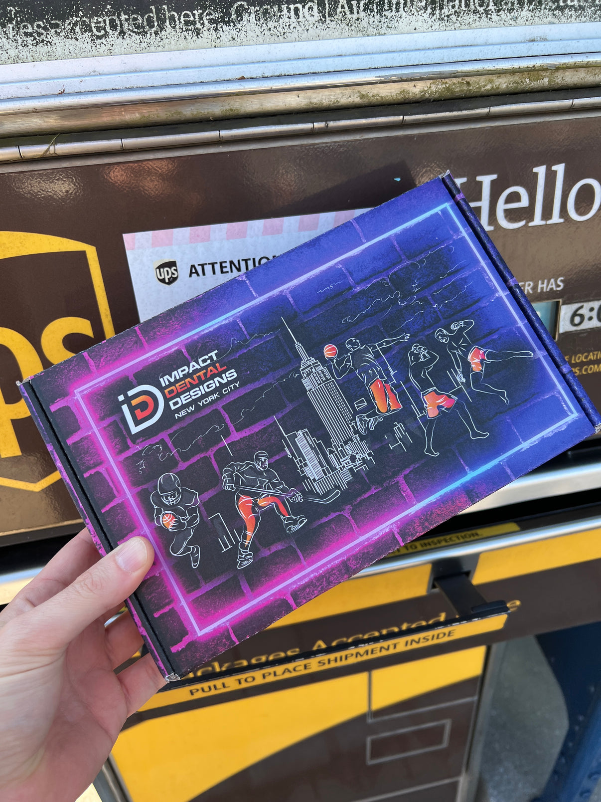 Hand holding an Impact Dental Designs package in front of a UPS drop box.
